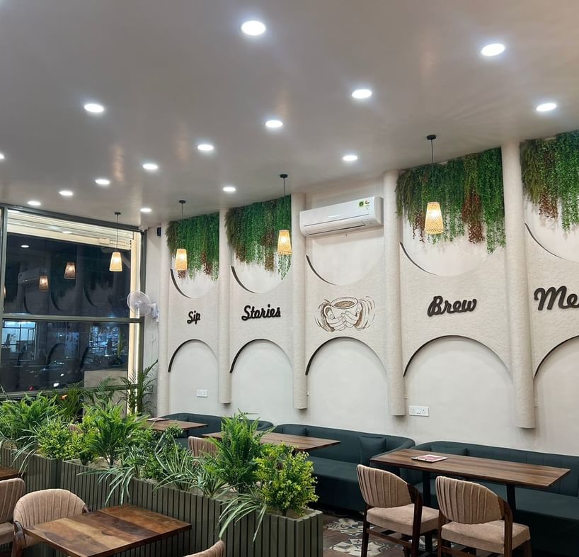 Aesthetic interior of Kafeholic Mansarovar in Jaipur featuring cozy seating, indoor plants, wall art reading ‘Sip Stories Brew Memories,’ warm hanging lights and a modern café ambience